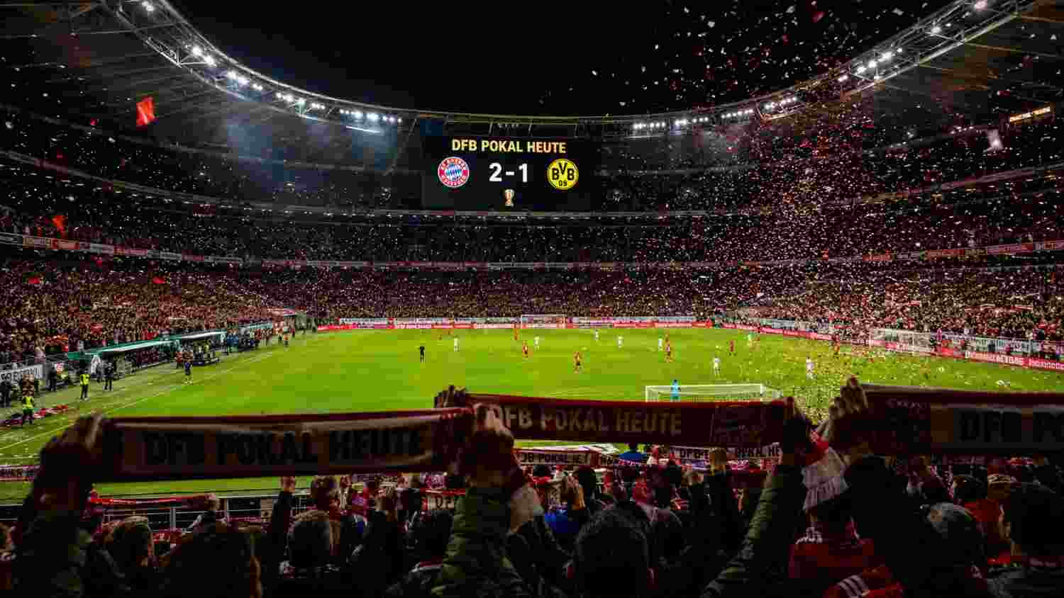 DFB Pokal heute live im Stadion: Fans halten Schals hoch, Anzeigetafel zeigt 2-1 Bayern gegen BVB bei Konfetti-Regen